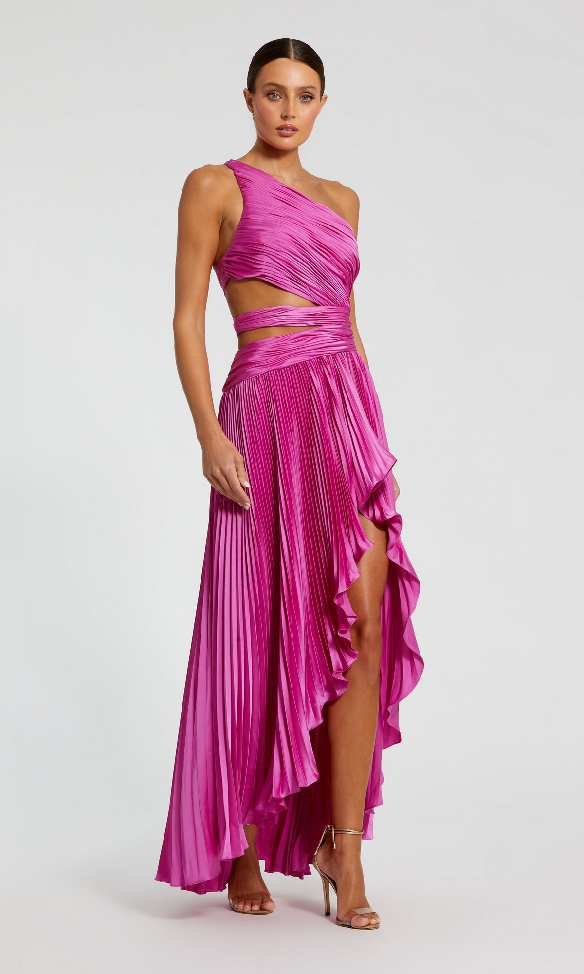 A woman in a bright pink Mac Duggal 49837 High-Low Pleated Formal Dress with a cut-out one-shoulder design and high slit, paired with silver heels, stands against a plain background.
